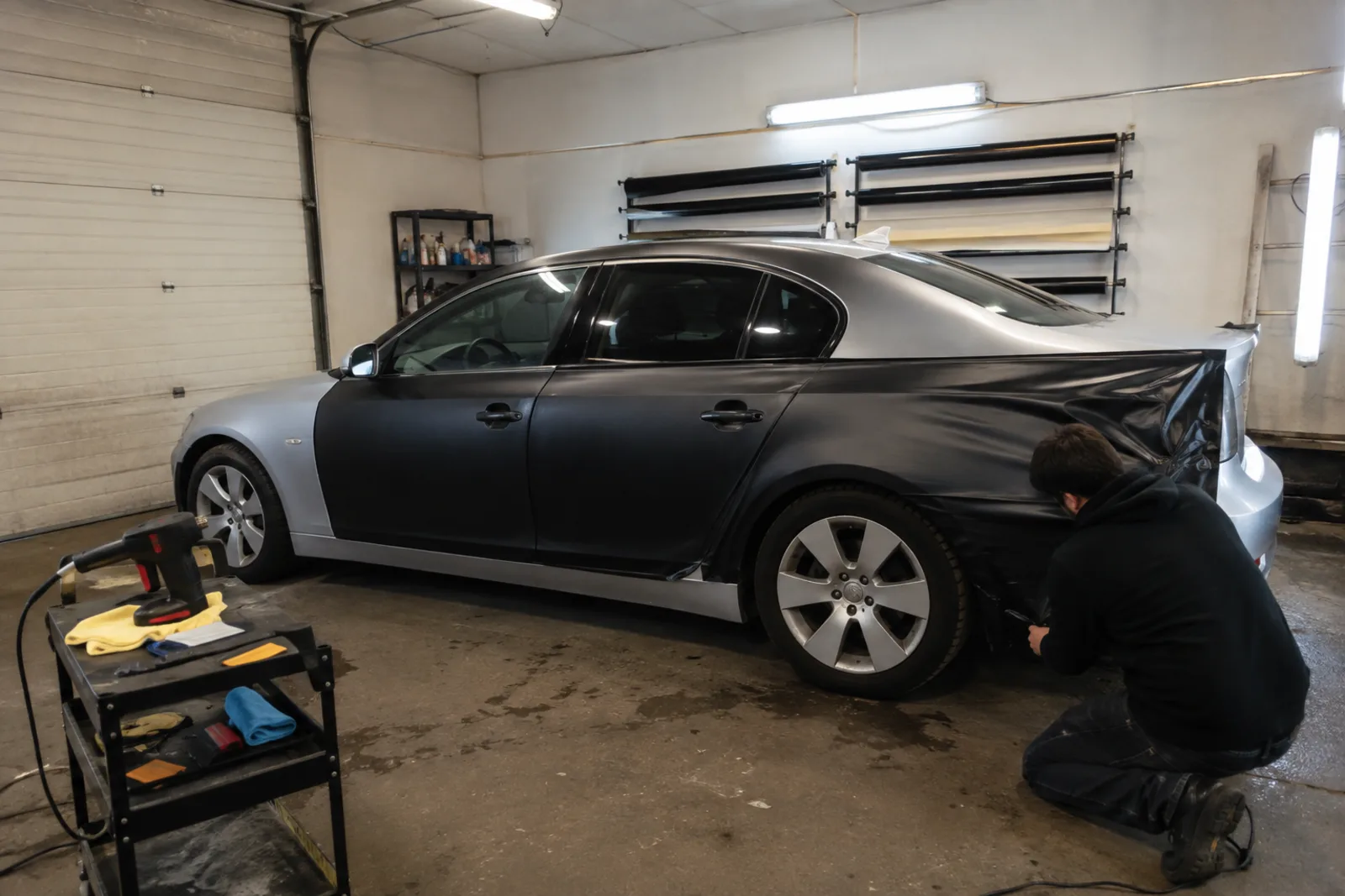 Silver sedan mid-wrap in a small garage workshop with vinyl tools on a cart